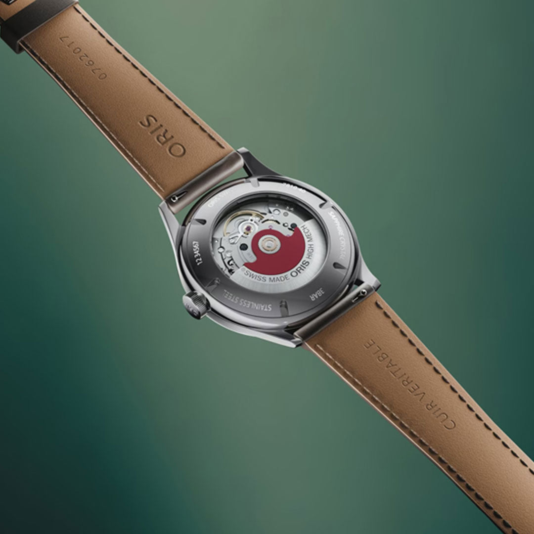 A close-up of the ORIS Artelier Date Calibre 733-1 Automatic 38mm Watch, showcasing its transparent case back with visible mechanical movement, brown leather strap, and stainless steel case against a green background.