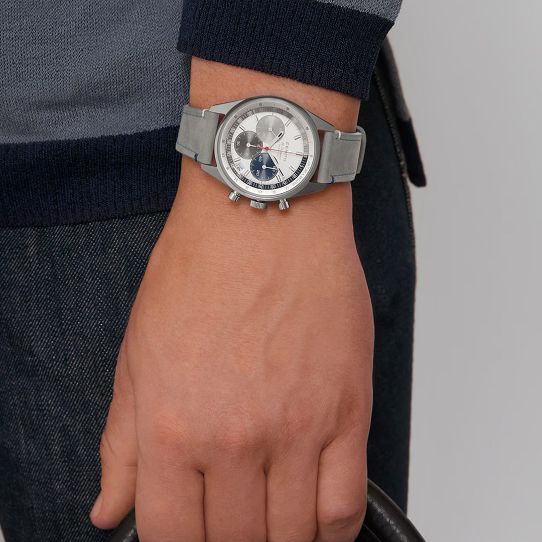 Wearing a Zenith Chronomaster Original El Primero 3600 38mm watch with a grey strap and three sub-dials, a person in a grey and blue sweater and dark pants holds a black object in their hand.