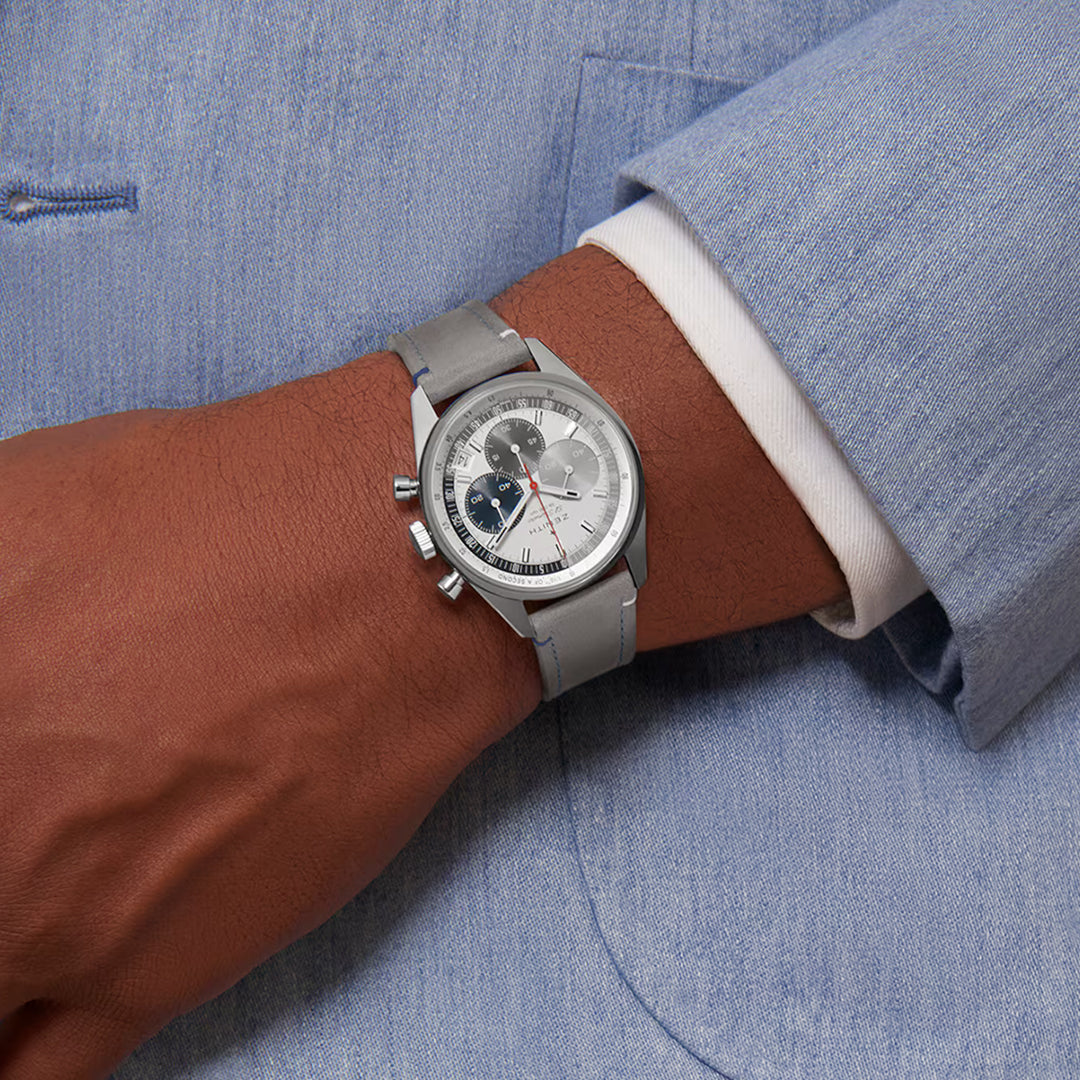 A person wears a light blue suit, highlighting the ZENITH Chronomaster Original El Primero 3600 38mm watch by Zenith. The silver wristwatch features a leather strap, white dial with blue and black subdials, and silver accents, showcasing its elegant high-frequency chronograph design.