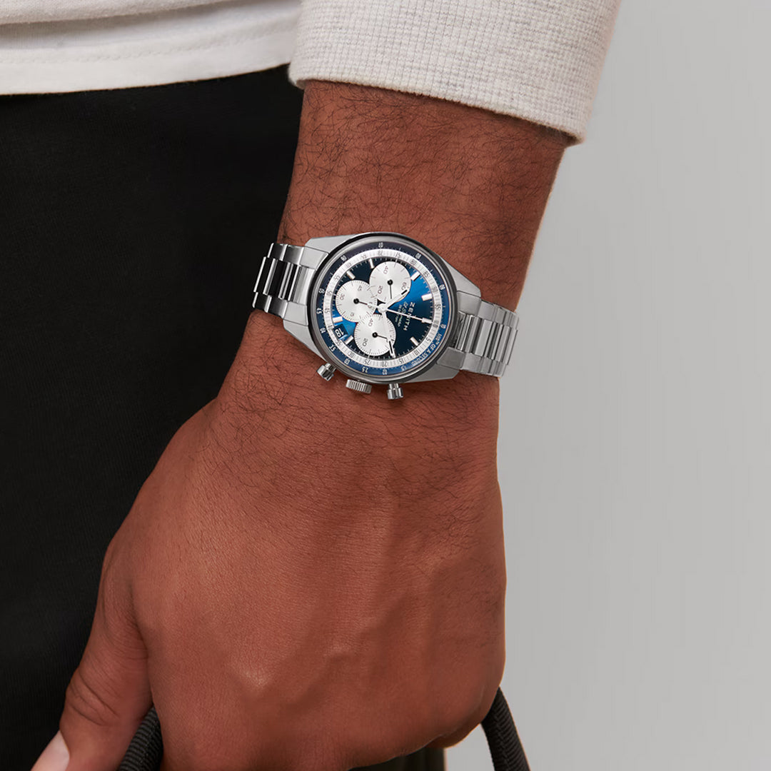 A person in a cream long-sleeve shirt and dark pants wears the ZENITH Chronomaster EP Original El Primero 3600 38mm Watch featuring a high-frequency chronograph with a blue face and silver sub-dials. They also carry a black bag.