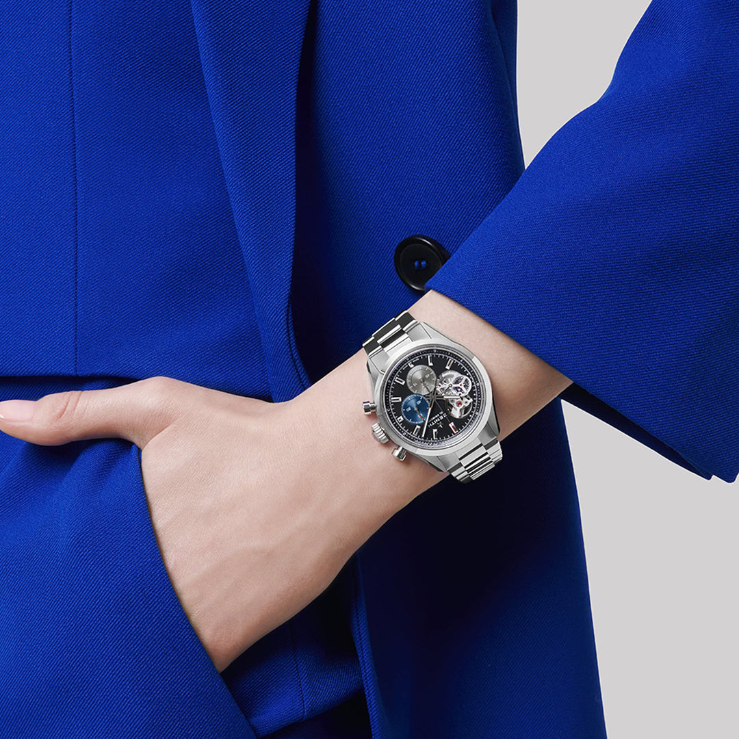 A person in a vibrant blue suit wears the Zenith Chronomaster Open El Primero 3604 on their left wrist. This watch has an automatic chronograph, a black face, intricate details inside its steel case, and a silver finish. Their hand is tucked into their pocket.