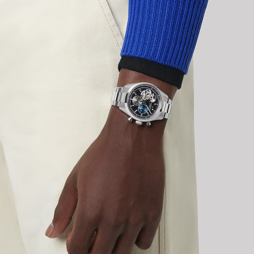 A person in a blue sweater and beige pants models the elegant Zenith Chronomaster Open El Primero 3604 wristwatch with its intricate dial, sophisticated silver tone, and steel case on their left wrist, all set against a plain gray background.