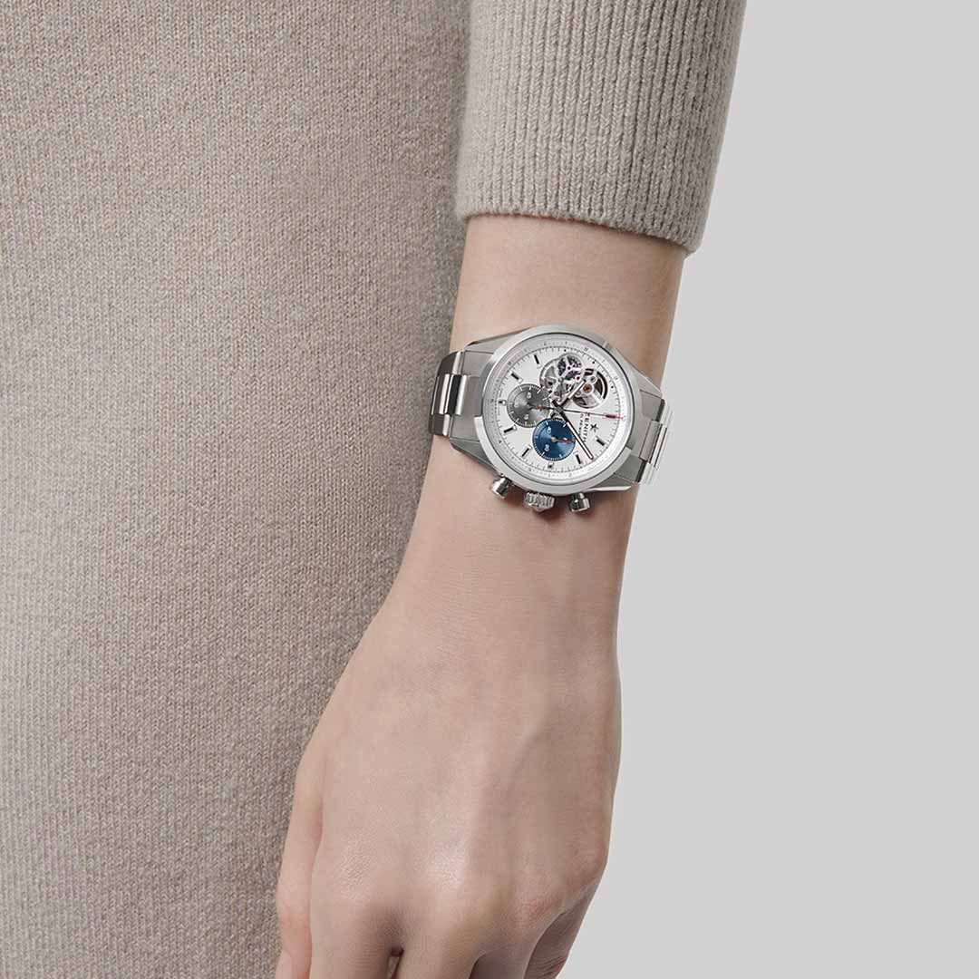 A person wearing a beige long-sleeve top displays the ZENITH Chronomaster Open El Primero 3604 Automatic 39.5mm Watch, featuring an intricate open dial that showcases the renowned El Primero movement with gears and blue accents. The focus is on the Zenith watch against a light gray background.