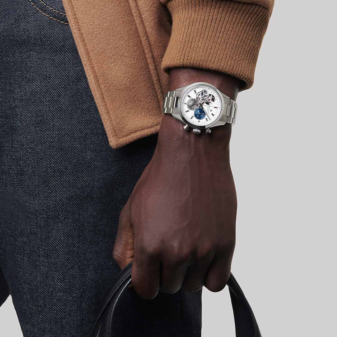 A person sporting a Zenith ZENITH Chronomaster Open El Primero 3604 Automatic 39.5mm watch, which reveals the intricate gears of the El Primero movement, is dressed in a tan jacket and dark jeans while holding a black item against a plain gray background.