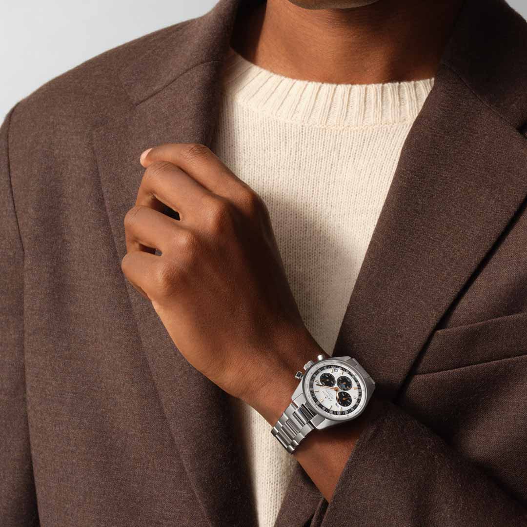 A person in a brown coat over a cream sweater showcases the ZENITH Chronomaster Original El Primero 3610 Automatic 38mm Watch by Zenith, highlighting its silver casing and black-and-white dial. The timepiece is prominently displayed on the left wrist, while the right hand adjusts the coat.