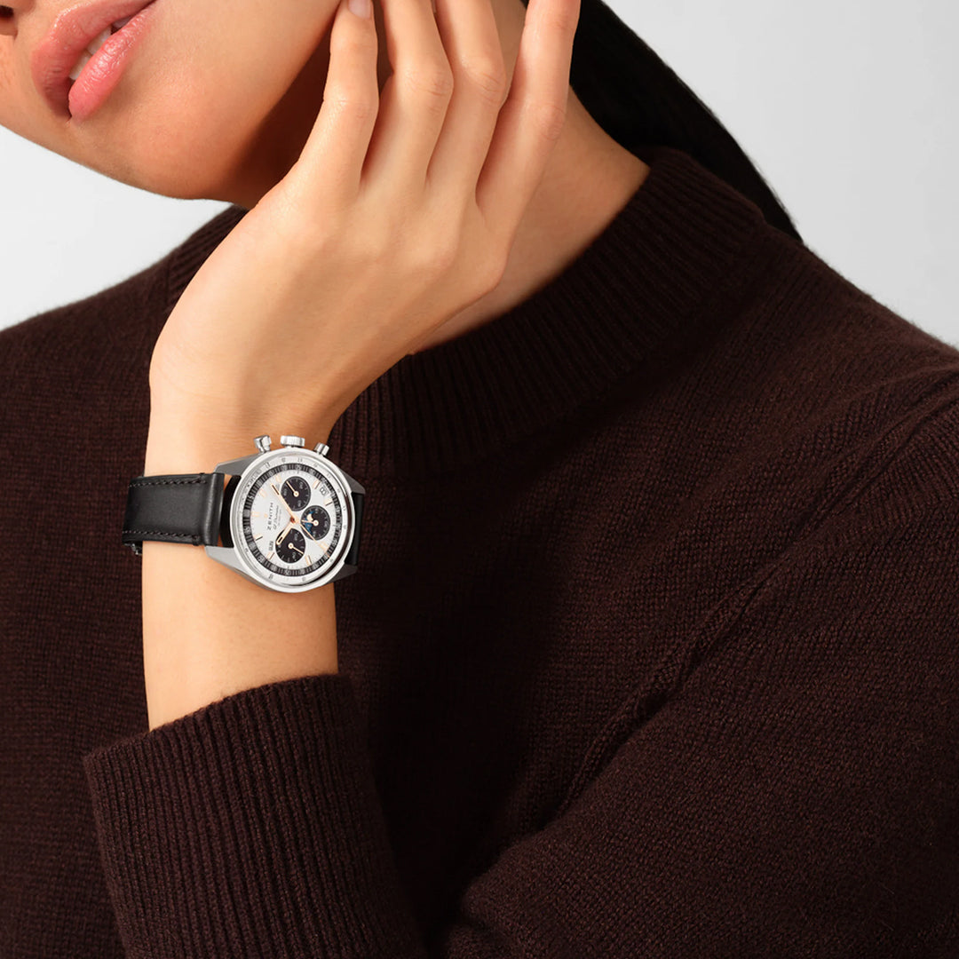 A person in a dark brown sweater wearing a ZENITH Chronomaster Original El Primero 3600 38mm Watch with a black leather strap rests their hand, adorned with the high-frequency chronograph, under their chin against a plain light gray background.