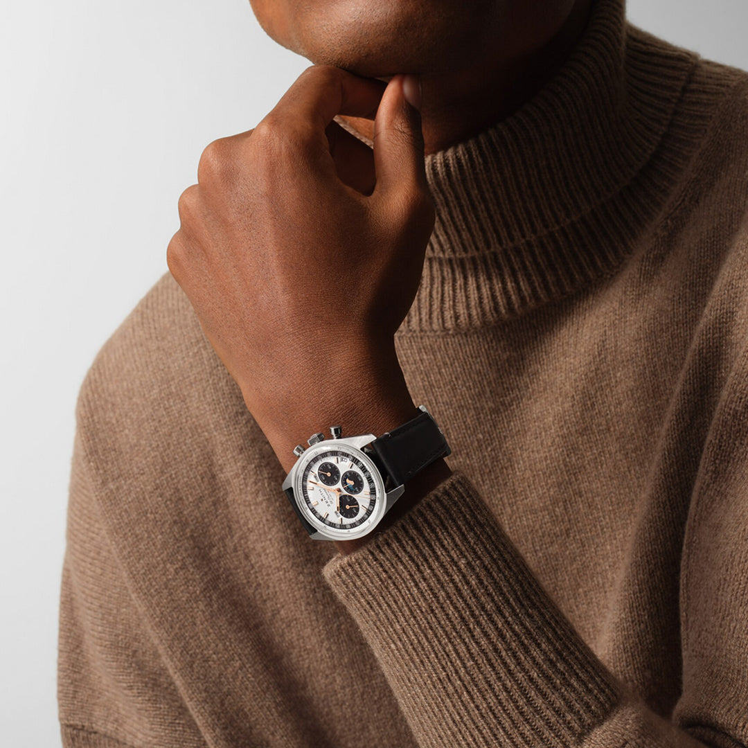 A person wearing a brown turtleneck rests their hand on their chin, showcasing a ZENITH Chronomaster Original El Primero 3600 38mm Watch. The silver timepiece elegantly features a black band and white face with sub-dials, embodying high-frequency chronograph movement against a plain background.