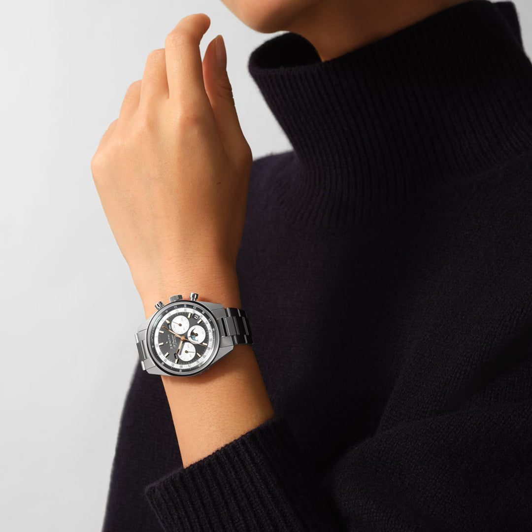 Against a white backdrop, a sleek silver ZENITH Chronomaster Original watch is worn by someone in a black turtleneck. It features three subdials on its black dial, echoing the precision and style of the El Primero 3610 movement.