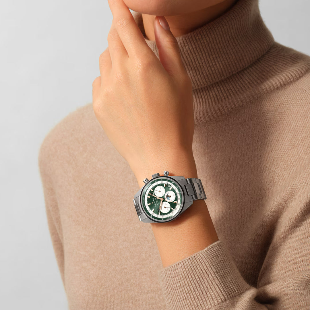 A person wearing a beige turtleneck poses thoughtfully with a hand near their chin, showcasing the Zenith Chronomaster Original El Primero 3610 watch. The timepiece features a green and white face, metal band, triple calendar, and three subdials.