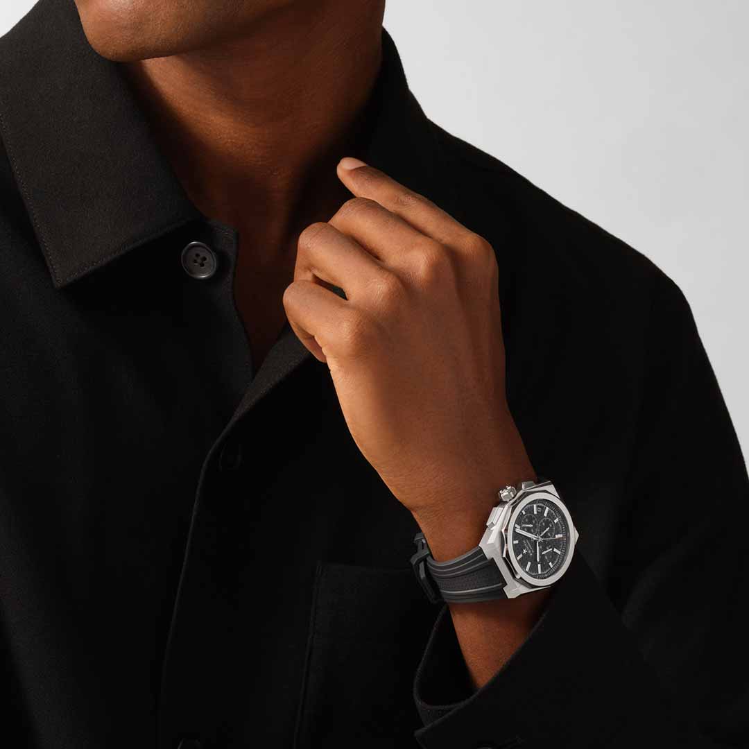 Person wearing a black shirt and a stylish Zenith DEFY Skyline Chronograph El Primero 3600 Automatic 42mm Watch with a black band and silver casing. Their hand is raised near their collar, exuding elegance. The watch's high-frequency movement adds an extra layer of sophistication to the overall image.