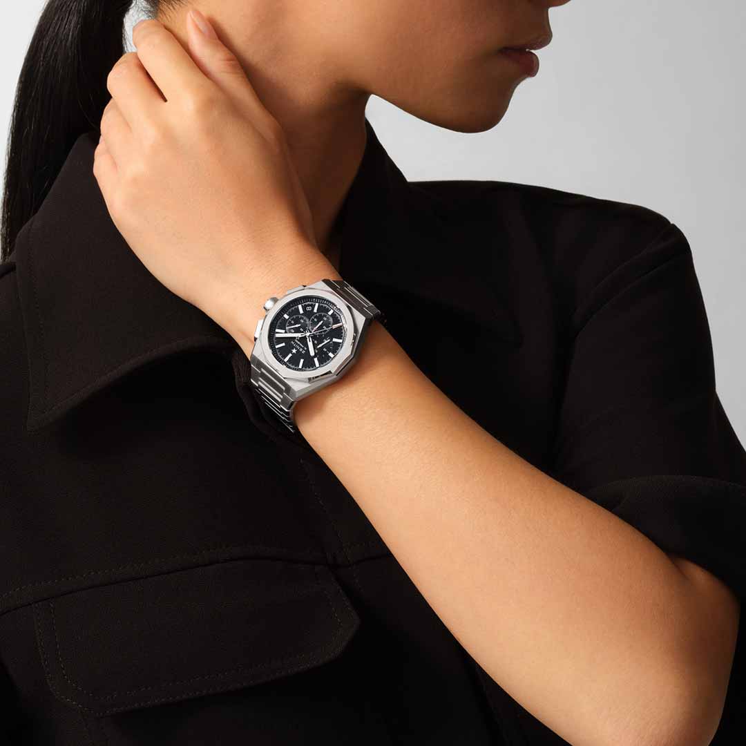 A person wearing a sleek black shirt adjusts a stylish Zenith ZENITH DEFY Skyline Chronograph El Primero 3600 Automatic 42mm Watch with a round black face on their wrist. The watch features chronograph dials and a metallic strap. The neutral background emphasizes the elegance of its high-frequency movement.