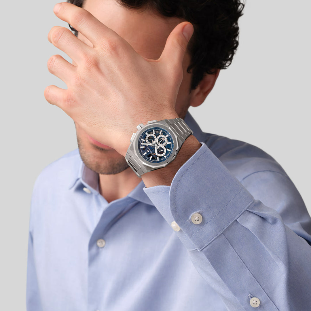 A person in a light blue shirt covers their face with one hand, showcasing a Zenith ZENITH Defy Skyline Chronograph Skeleton El Primero 3600SK 42mm Watch with a detailed dark watch face. The background is plain gray.