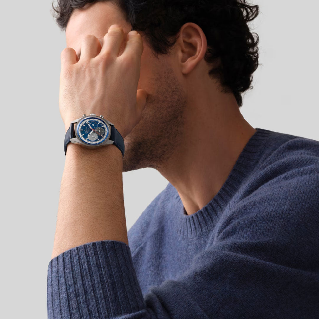 A person with short, curly hair in a blue sweater partially covers their face with their left hand, showing a Zenith Chronomaster Original El Primero 3610 38mm watch with a blue dial on their wrist.