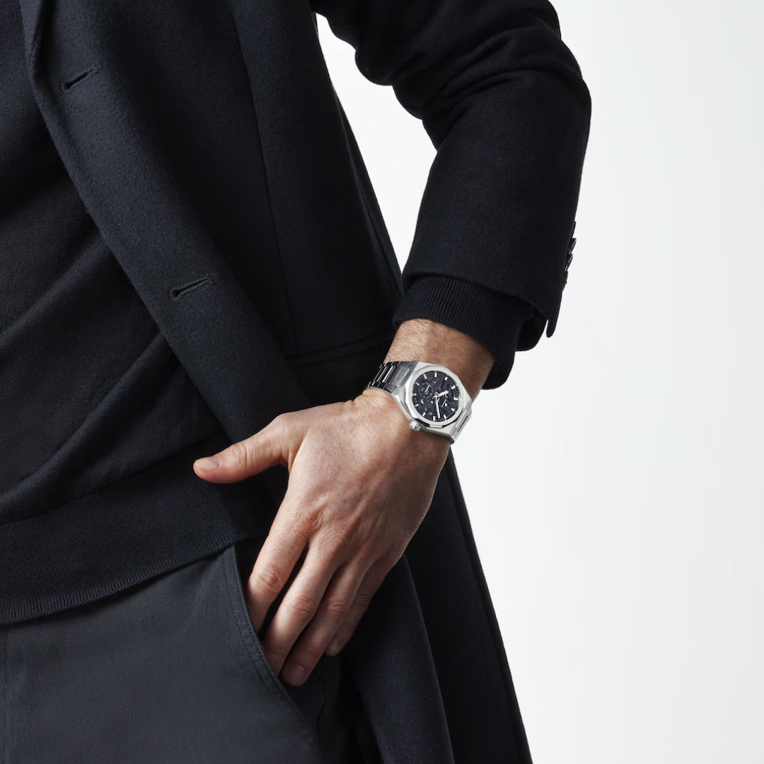 A person stands against a plain backdrop in a dark coat, hand in pocket, showcasing the ZENITH Defy Skyline Skeleton El Primero 3620 SK Automatic 41mm Watch, featuring an interchangeable strap and black face.