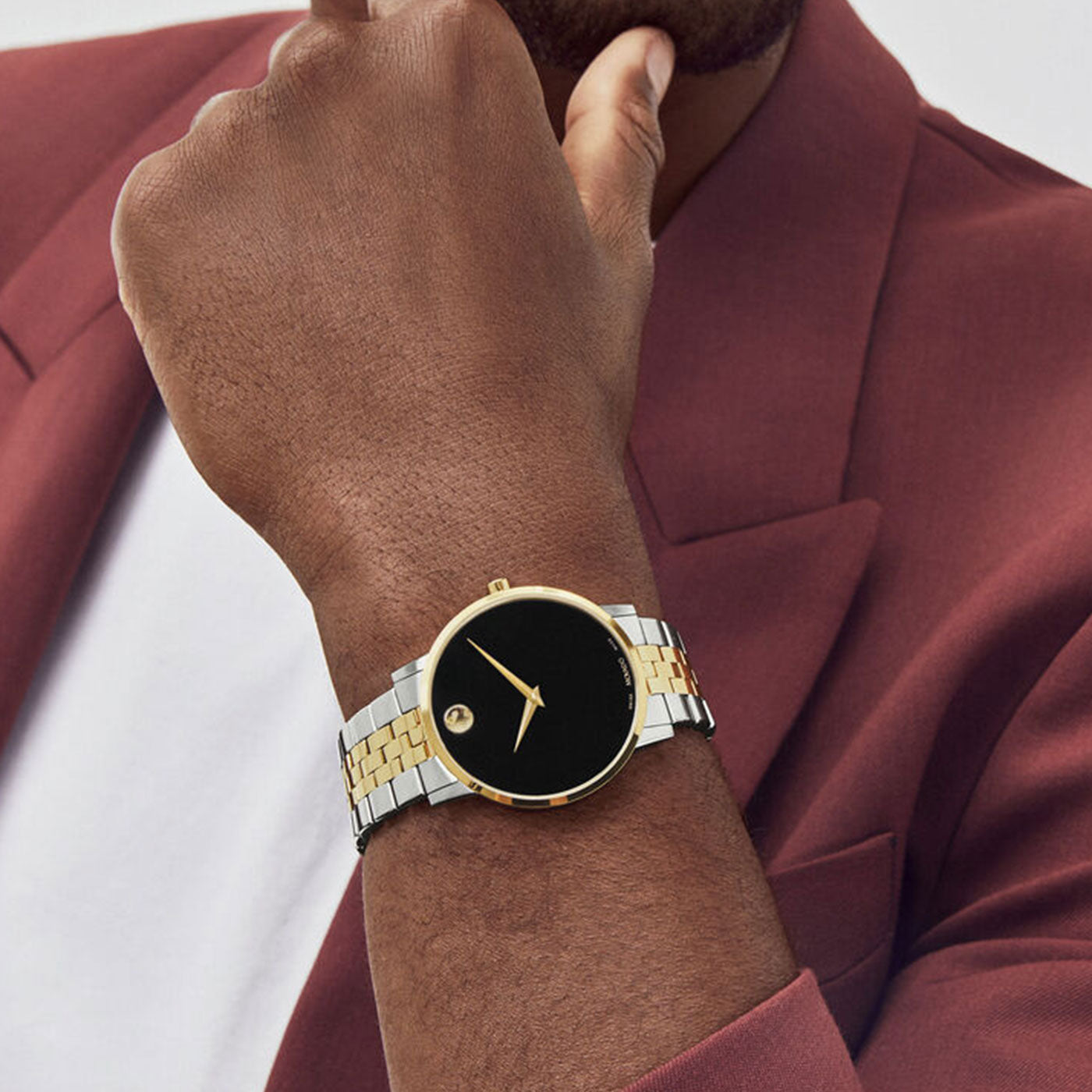 A person in a maroon blazer and white shirt is showcasing the Movado Museum Classic Quartz 42mm watch. This luxury timepiece from Movado features a sophisticated combination of gold and silver strap, stainless steel detailing, with a black face and yellow gold PVD rim, exuding an elegant look. The individual is resting their chin on their hand.