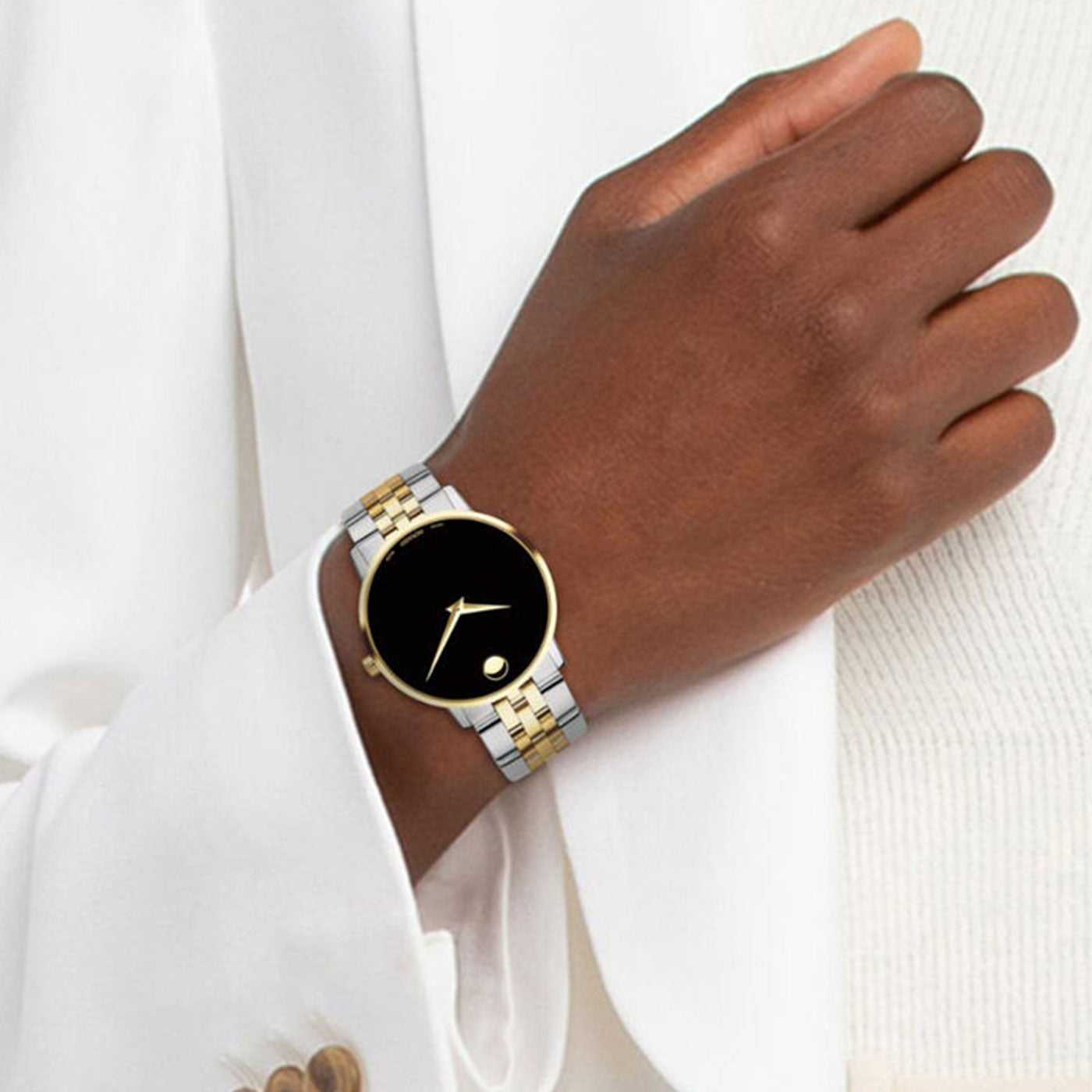 A person wearing a stylish Movado Museum Classic Quartz 42mm watch, characterized by its round black face, gold accents, and two-tone stainless steel band. The individual is dressed in a white outfit.