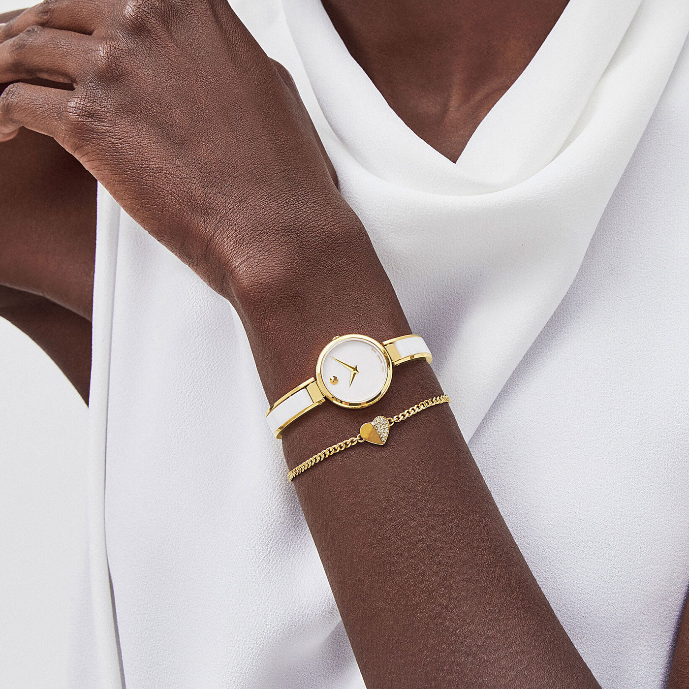 A person wearing a white sleeveless top showcases the chic Movado Moda Quartz 24mm Watch, elegantly paired with a gold bracelet adorned with a dainty heart charm.