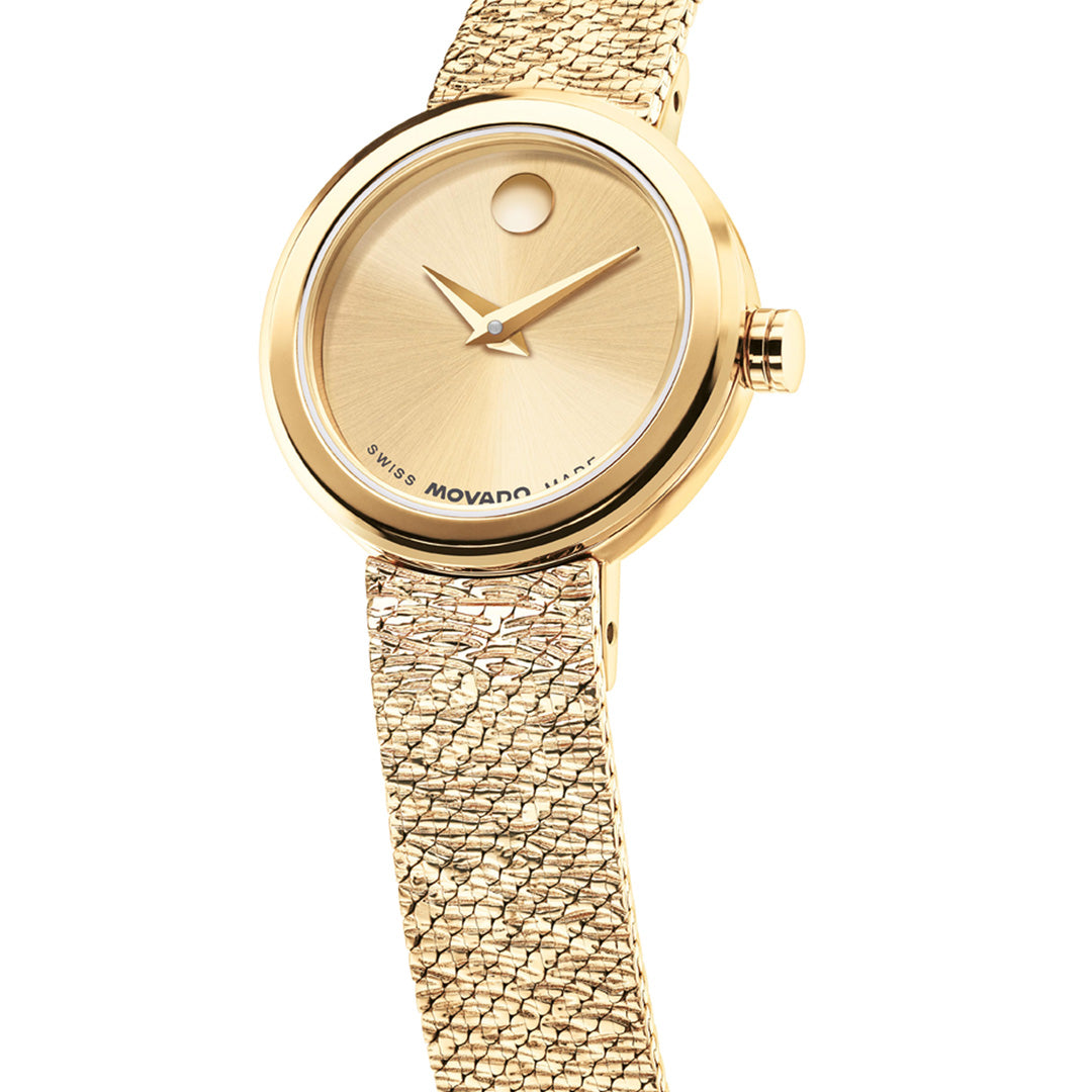 The Movado Veluran Mini Quartz 21mm Watch by Movado features a round, minimalist yellow gold face accented with a single dot at 12 o’clock and is complemented by a textured Milanese mesh bracelet, elegantly angled against a white background.