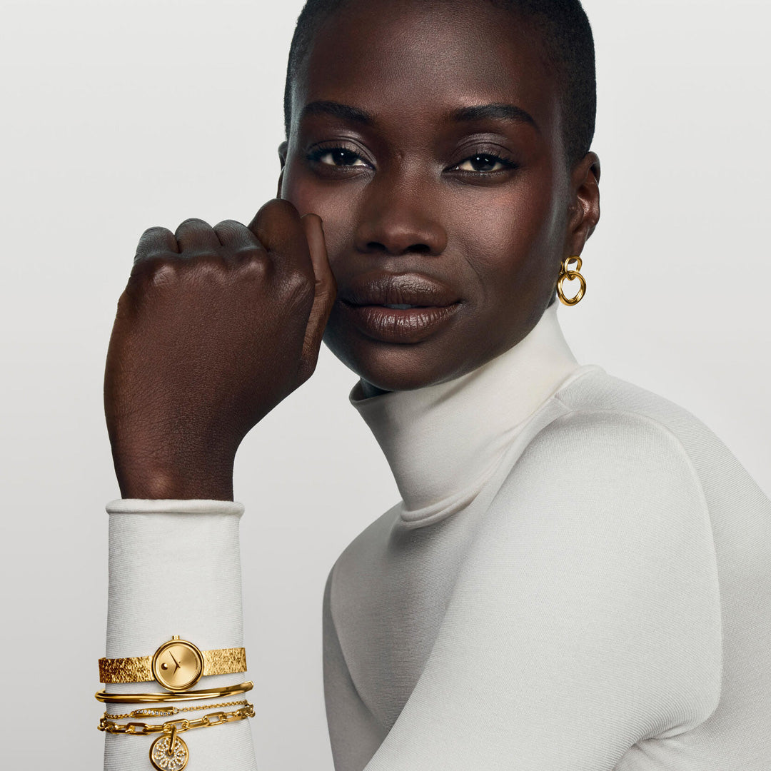 A woman in a white turtleneck poses with her hand under her chin, wearing gold hoop earrings, several gold bracelets, and the Movado Veluran Mini Quartz 21mm Watch featuring a yellow gold PVD case. The background is plain and gray.