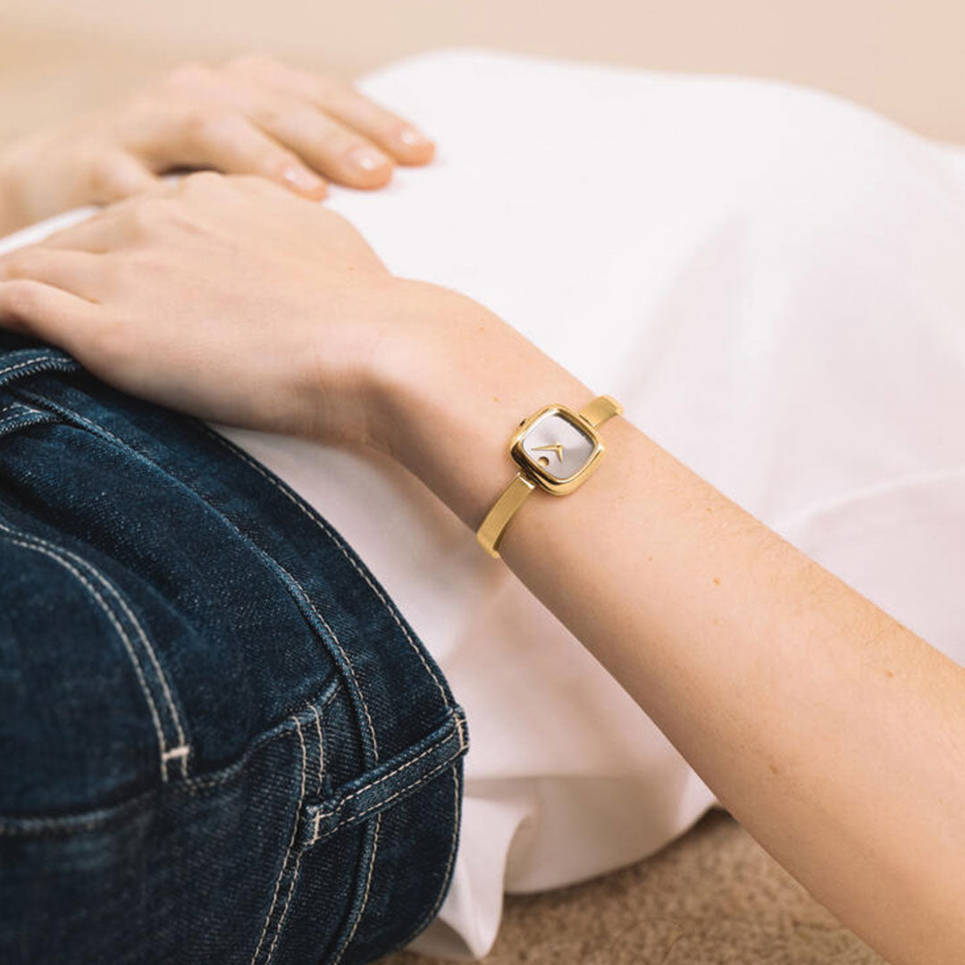 A person in blue jeans and a white shirt lies down, showing a close-up of their wrist wearing the Movado Museum Bangle Quartz 21.5mm Watch—a minimalist gold square-face design from Movado, echoing classic elegance.