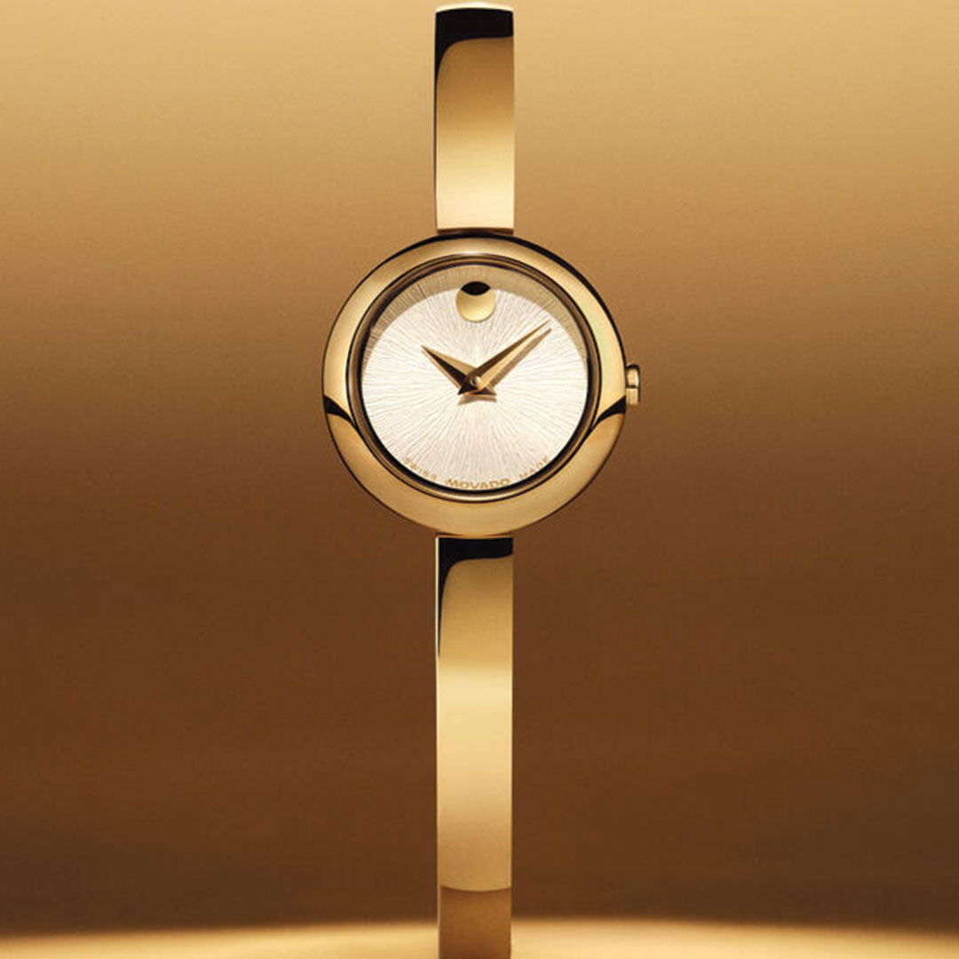 The Movado Museum Bangle Quartz 21.5mm Watch, featuring a gold tone and signature single dot at 12 o’clock, is shown against a matching gold background. Its jewelry-inspired band highlights the sleek, modern design by Movado.