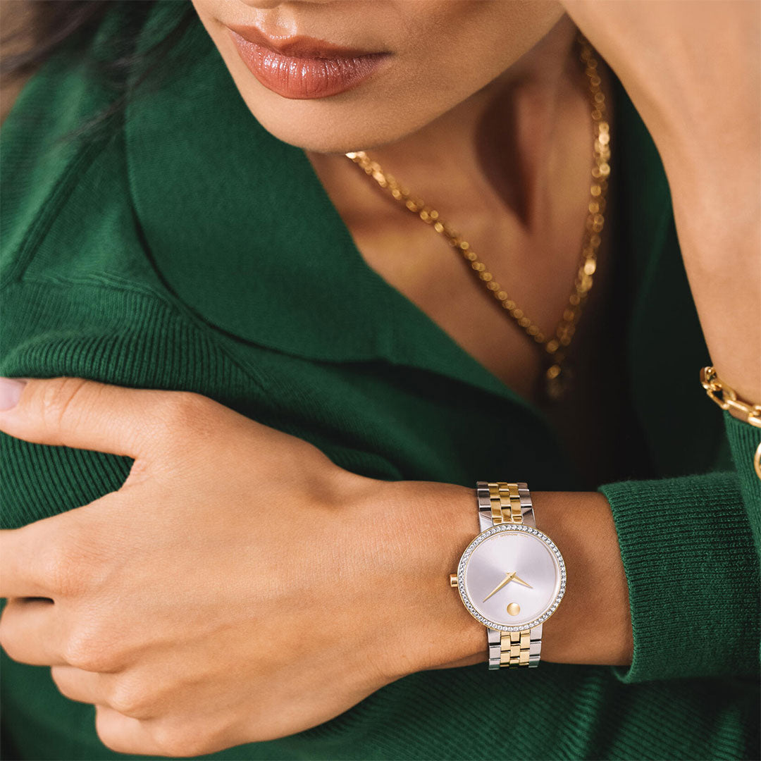 A woman wearing a green sweater and gold jewelry rests her arm, showcasing the Movado Museum Classic Quartz 29.5mm Watch by Movado, featuring a two-tone design with a silver white sunray dial and gold accents.