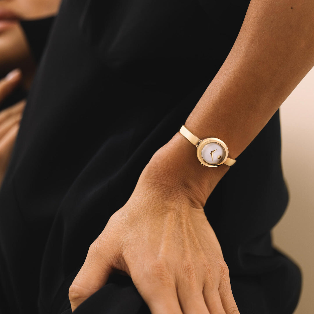 Close-up of a person wearing the Movado Museum Bangle Quartz 21.5mm x 25.5mm Watch by Movado—a minimalist, jewelry-inspired gold wristwatch with a round white face—dressed in a black outfit. The person’s hand rests casually near their hip, prominently displaying the elegant watch.
