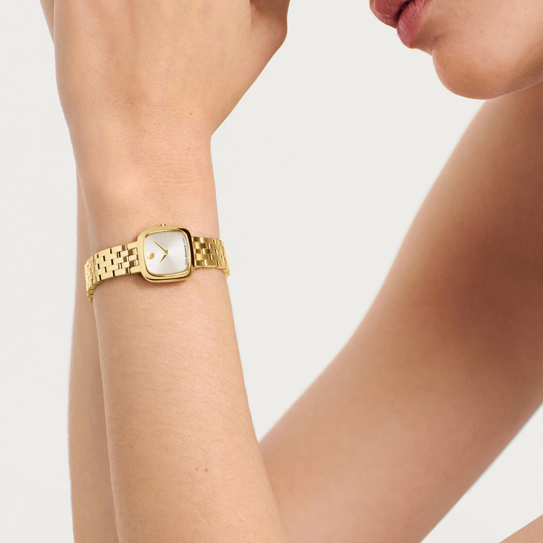 A person wearing the Movado Museum Classic Quartz 22mm x 22mm watch by Movado, featuring a yellow gold tone and a square white dial, displayed on their left wrist against a neutral background.