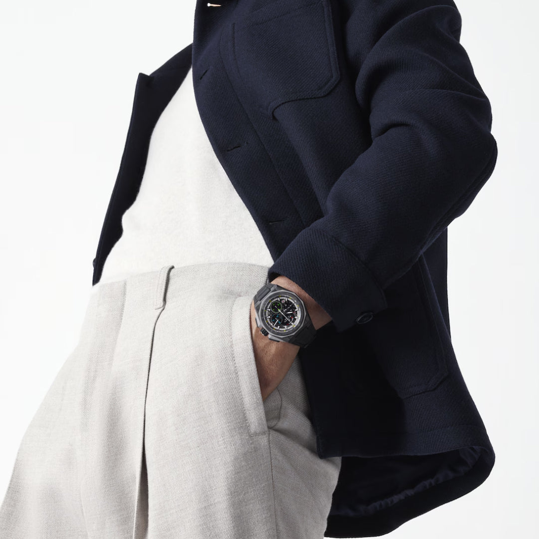 A person wears a black jacket and white shirt with beige trousers. On their left wrist is the striking Zenith Defy Extreme Carbon El Primero 21 Automatic 45mm Watch. The background is plain and light-colored.