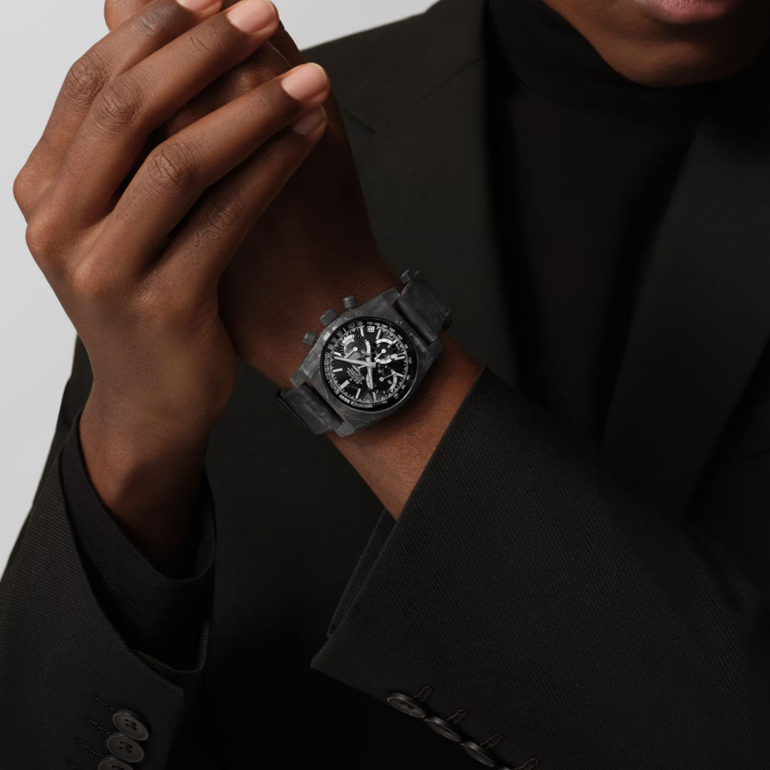 A person in a black suit and turtleneck showcases the Zenith Chronomaster Revival El Primero 400 37mm watch, featuring a detailed black chronograph dial with white accents, holding one hand near the timepiece.
