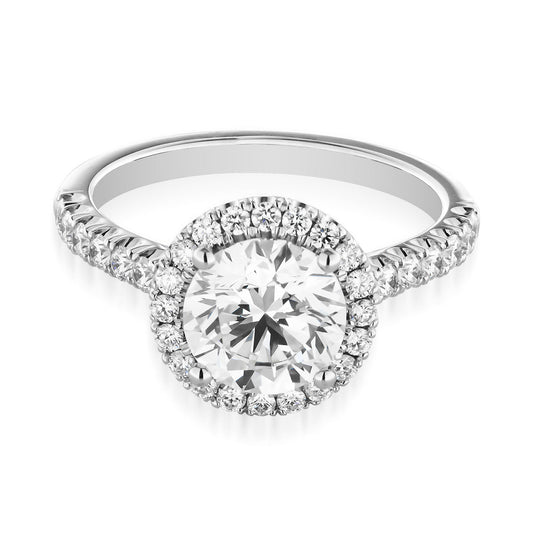 The Raffi&Co.® Signature 14K White Gold Round Brilliant Halo Diamond Engagement Ring showcases a stunning centerpiece with a large round brilliant diamond, encircled by a halo of smaller diamonds. The band, embellished with additional diamonds, enhances the sparkle and elegance of this exquisite design.