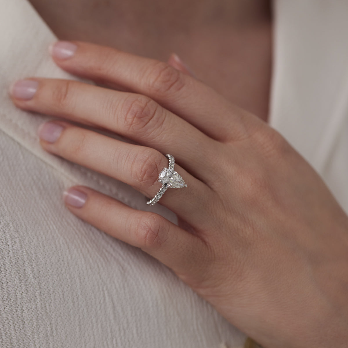 A hand with a pale skin tone rests on white fabric, adorned with the Raffi&Co.® 14K White Gold Pear Hidden Halo Diamond Engagement Ring. This exquisite piece, crafted by Raffi&Co, features a 1.70 carat pear-shaped diamond and a delicate band accented by smaller diamonds. The nails are neatly manicured with a natural finish, showcasing elegance and simplicity.