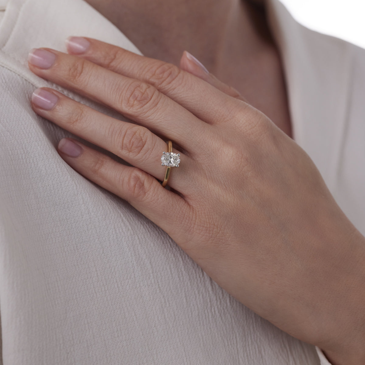 A person adorns their left ring finger with a Raffi&Co.® 14K Yellow Gold Oval Solitaire Diamond Engagement Ring. Their hand rests gracefully on the upper chest, over a cream-colored garment, showcasing well-manicured nails.