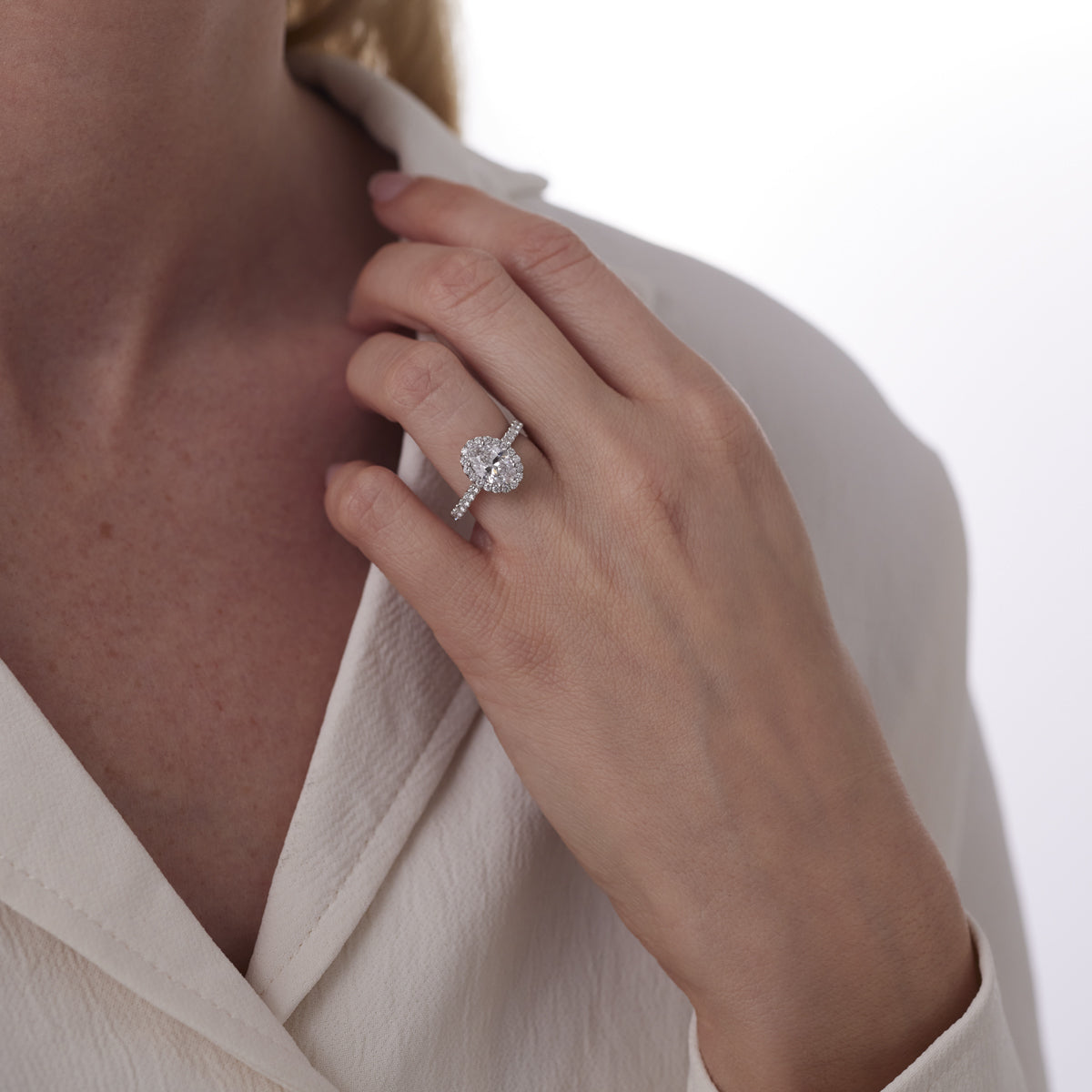 A person wearing a white shirt is showcasing a Raffi&Co.® 14K White Gold Oval with Round Brilliant Halo Diamond Engagement Ring on their left hand. The GIA-certified ring, by Raffi&Co, features an oval central diamond stone surrounded by smaller diamonds adorning the band of 14K white gold. The background is softly blurred.