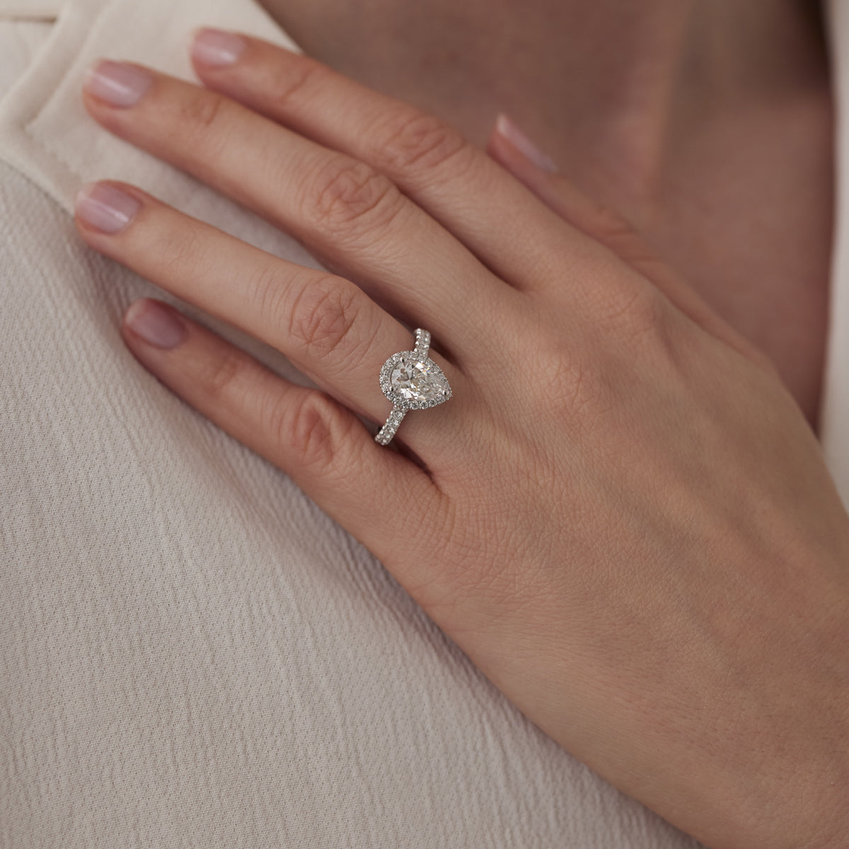 A close-up of a hand wearing the Raffi&Co.® 14K White Gold Pear Halo Diamond Engagement Ring features the dazzling gem on a detailed band, resting on light-colored fabric and accentuating well-manicured nails.