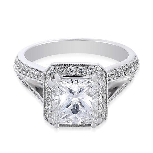 The Raffi&Co.® 14K White Gold Princess Cut Halo Diamond Engagement Ring features a prominent, square-cut diamond at its center, emulating the elegance of a princess cut. This striking centerpiece is surrounded by smaller diamonds set along the band, with intricate detailing and a split shank setting enhancing its sophisticated design.