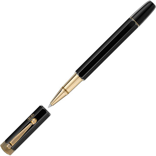 The Montblanc Heritage Egyptomania Special Edition Rollerball Pen, featuring a stylish black and gold hexagonal barrel adorned with Egyptian-style engravings, is displayed with its cap removed. Positioned diagonally, it showcases the elegant design from the Montblanc Heritage Collection. The cap lies beside the open pen, highlighting its gold details.