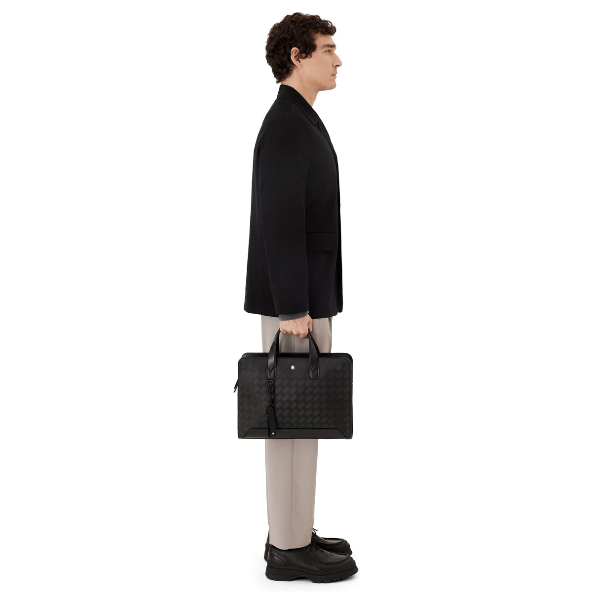 A man in a black jacket and beige pants stands in profile, holding the Montblanc Extreme 3.0 Black Leather Document Case. Wearing black shoes, he exudes elegance against the white background.