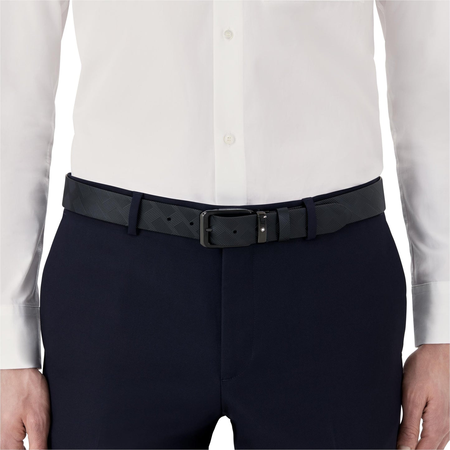 A person in a white dress shirt is wearing black formal pants paired with a Montblanc Extreme 3.0 Reversible Leather Belt. The belt showcases a subtle checkered pattern and is fastened with a rectangular metal buckle, capturing the waist area as the focal point of the image.