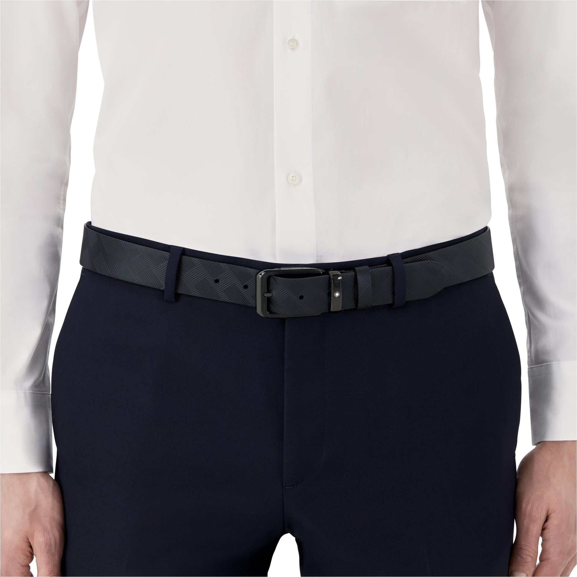 A person in a white dress shirt is wearing black formal pants paired with a Montblanc Extreme 3.0 Reversible Leather Belt. The belt showcases a subtle checkered pattern and is fastened with a rectangular metal buckle, capturing the waist area as the focal point of the image.