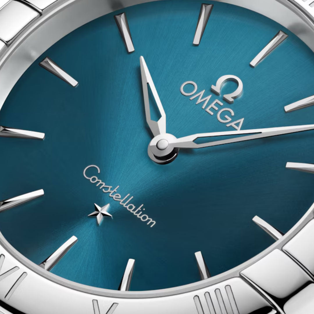 Close-up of the OMEGA Constellation Quartz 28mm Watch, featuring a peacock blue dial with silver hour markers, polished hands, and the Omega logo with "Constellation" text above a star on a stainless steel case.