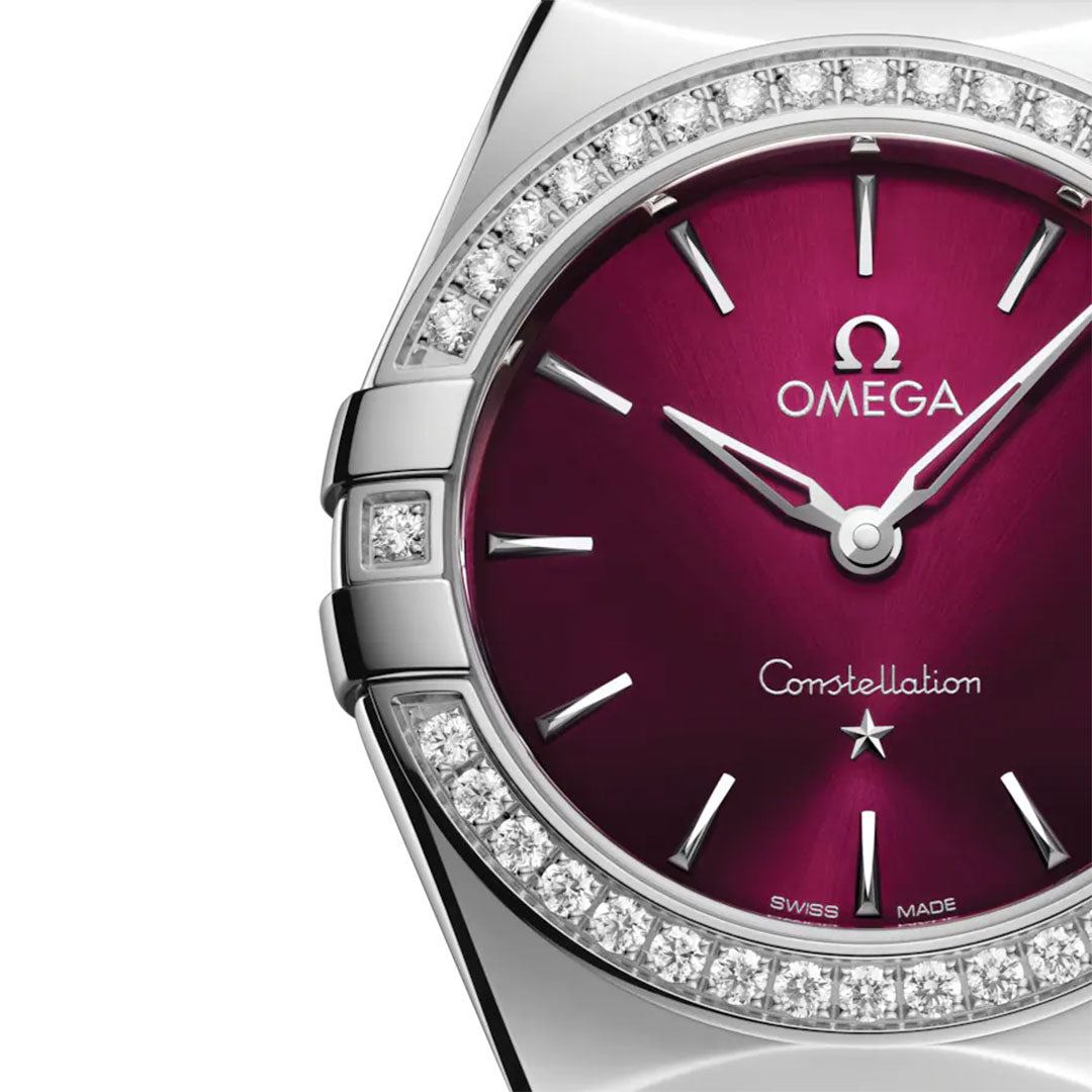 Close-up of the OMEGA Constellation Quartz 28mm Watch in stainless steel, displaying a deep red sunburst dial with silver hands and hour markers, a diamond-paved bezel, and visible Omega logo and "Constellation" on the dial.