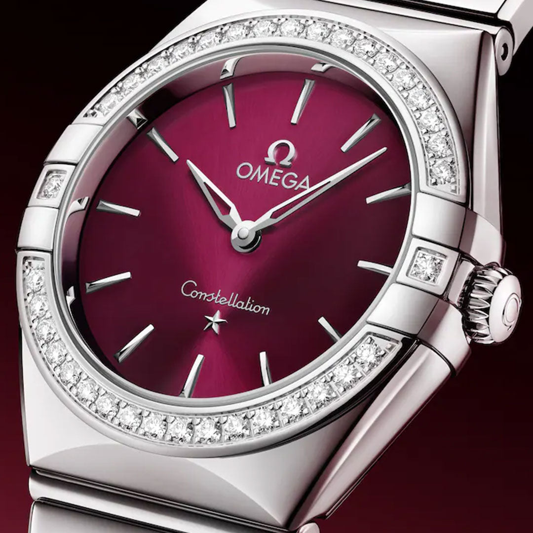 A close-up of the OMEGA Constellation Quartz 28mm Watch featuring a deep red dial, silver hour markers and hands, a diamond-paved bezel, and a sleek stainless steel case and bracelet for refined elegance.