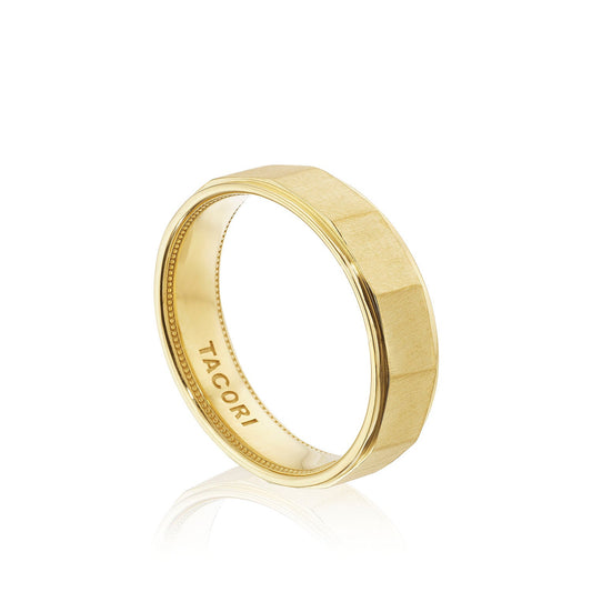 An 18K yellow gold men's wedding band with geometric facets and a satin finish stands upright on a reflective white surface. The word "TACORI" is elegantly engraved inside, highlighting the distinct Tacori crescent design subtly etched along the inner curve.