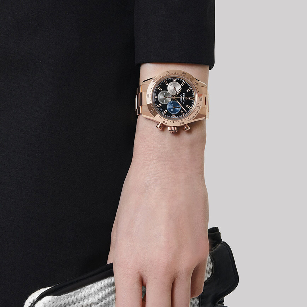 An individual flaunts a Zenith Chronomaster Sport El Primero 3600 41mm watch with a tricolour dial and chronographs. They clutch a textured black and white purse against a grey backdrop while dressed in an elegant black long-sleeve garment.