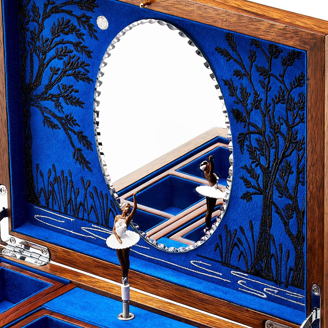 The WOLF 1834 Ballet Musical Jewellery Box by WOLF celebrates Swan Lake with a mirror and a ballerina in a white tutu. Its interior is lined with blue velvet, showcasing black embroidered trees and grass, and it features numerous compartments for ample storage of your treasures.