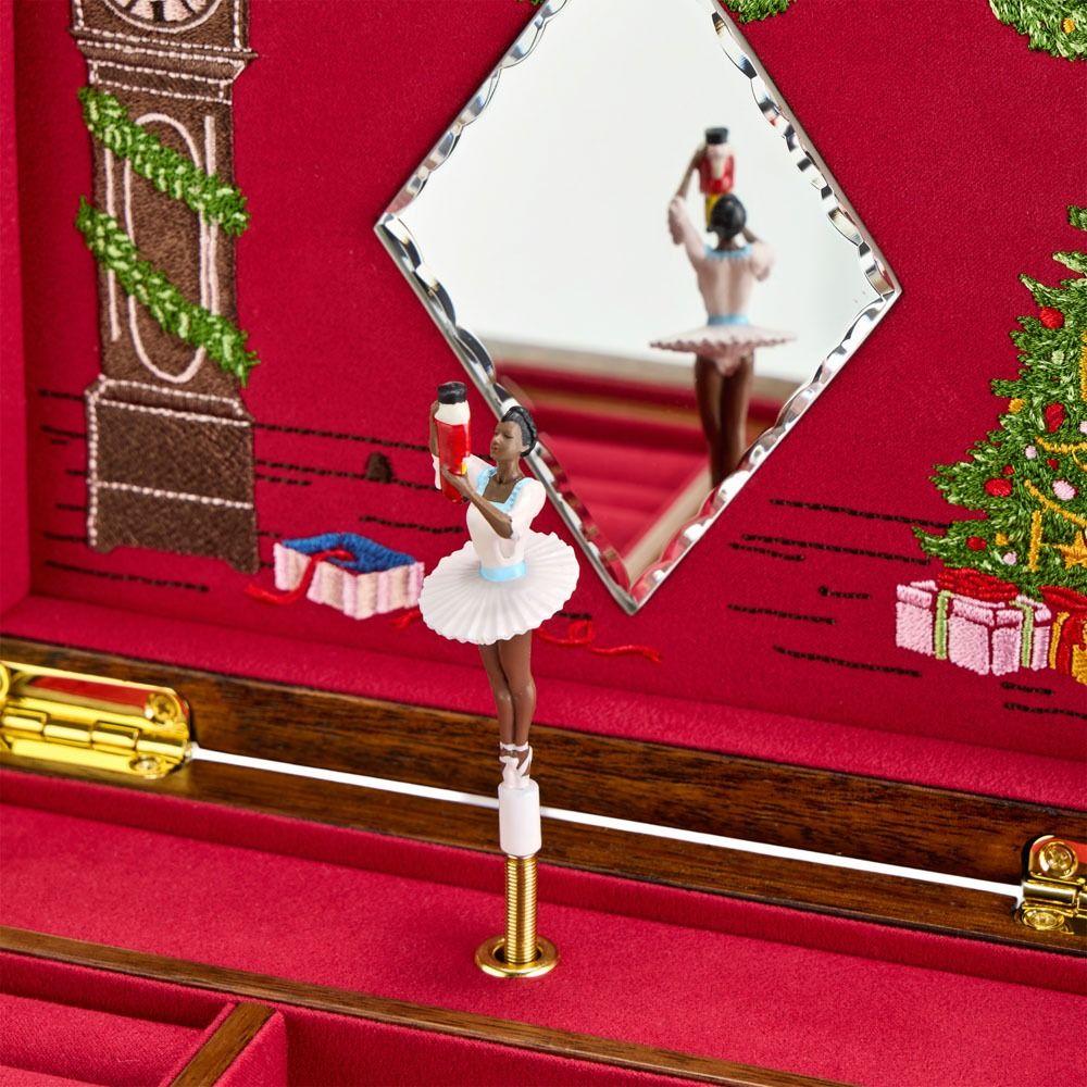 A ballerina figurine in a white tutu stands gracefully inside the WOLF 1834 Nutcracker Musical Box Jewelry Box Walnut, reflected in the interior mirror. Behind her, embroidered images of a clock tower, tree, and presents create a festive scene inspired by the Nutcracker ballet from WOLF.