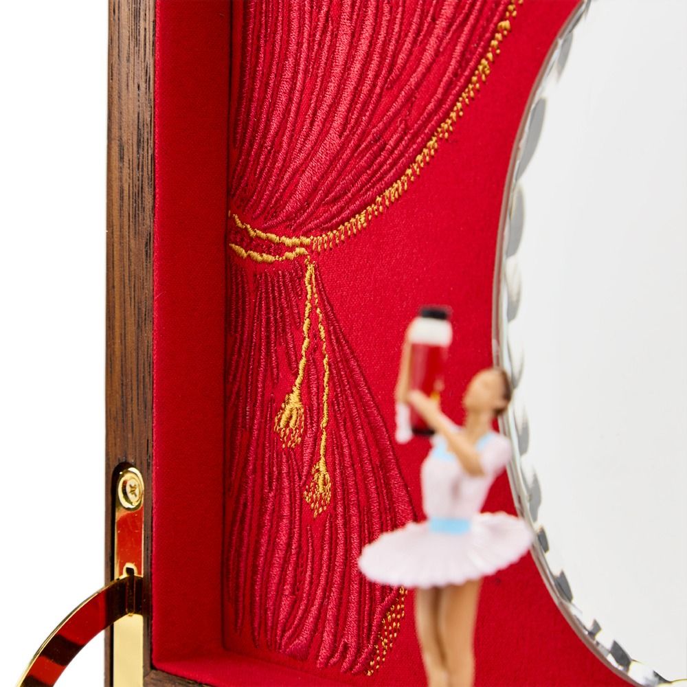 A close-up of a ballerina figurine, poised gracefully in front of an embroidered red curtain background and a decorative mirror inside the WOLF 1834 Nutcracker Musical Box Jewelry Box Walnut by WOLF.
