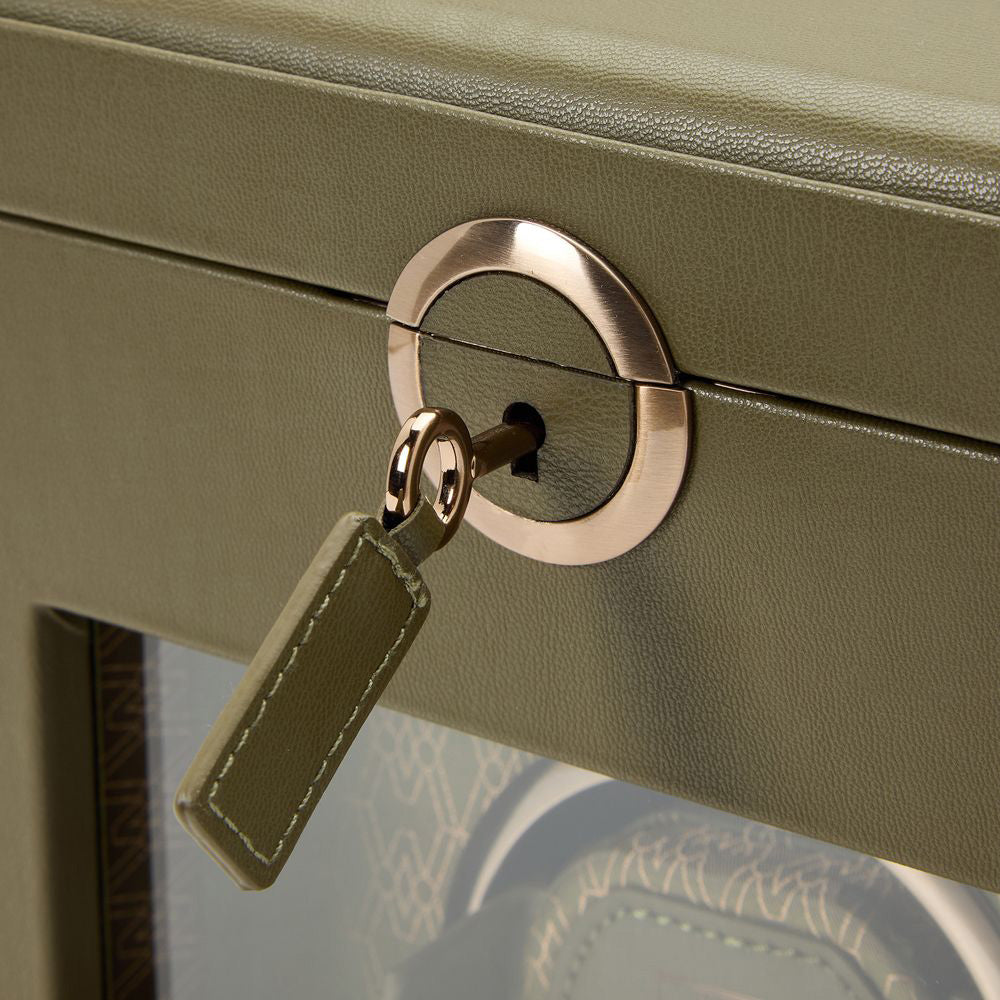 A detailed view of the WOLF 1834 Earth Single Watch Winder highlights its green leather exterior and distinctive gold circular lock, featuring a unique patented lock-in dynamic cuff. A key with a matching green leather tag is inserted into the lock, while the interior showcases a pattern suggesting luxury branding by WOLF.