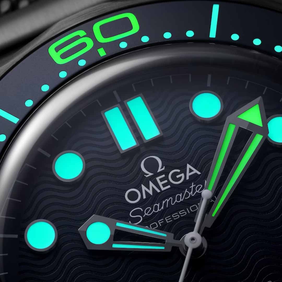 A close-up of the OMEGA Seamaster Diver 300M James Bond 60th Anniversary Co-Axial Master Chronometer 42mm Watch face showcases luminescent hour markers and hands. The vibrant green glow stands out against the dark background, evoking a classic James Bond feel while prominently featuring OMEGA’s name and logo.